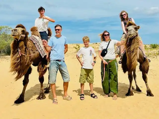 Family posing with two camels on a sand dune under a blue sky. Travel to Asia with kids